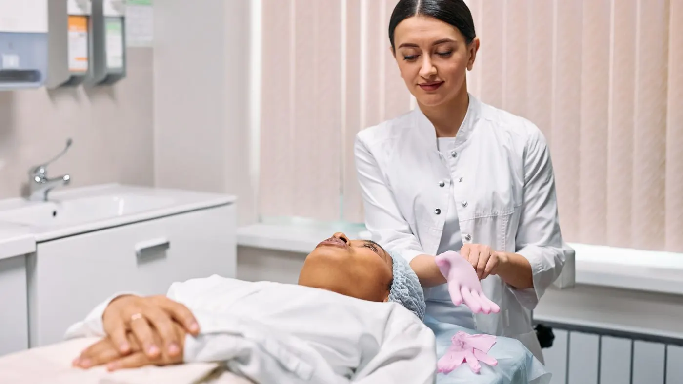 Médica con bata blanca que se pone guantes rosados junto a una paciente acostada en la camilla de un consultorio, antes de un procedimiento estético como la carboxiterapia