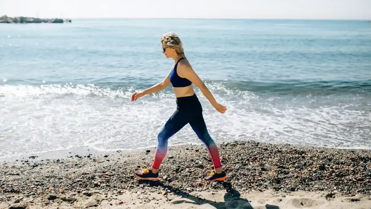 Mujer con ropa deportiva que camina a paso firme por la orilla de una playa de piedras y arena, el tipo de actividad sostenida con la que el cuerpo recurre a sus reservas de grasa